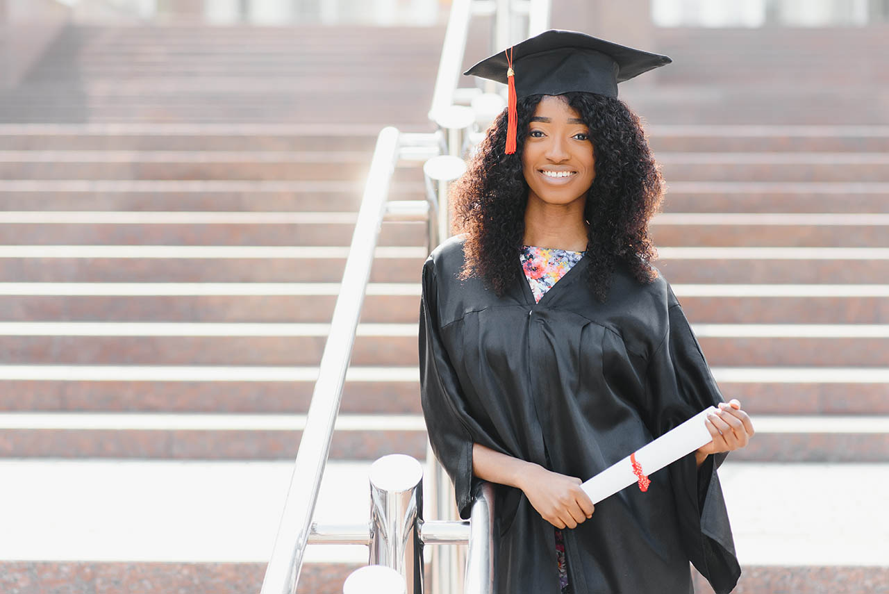 cheerful afro american female graduate standing in front of university building