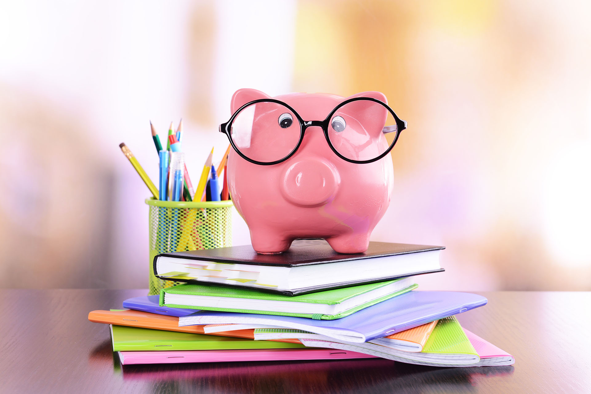 Piggy bank wearing glasses sits on a stack of notebooks and a planner next to a pencil cup filled with colored pencils, pens, and pencils on a wooden surface.