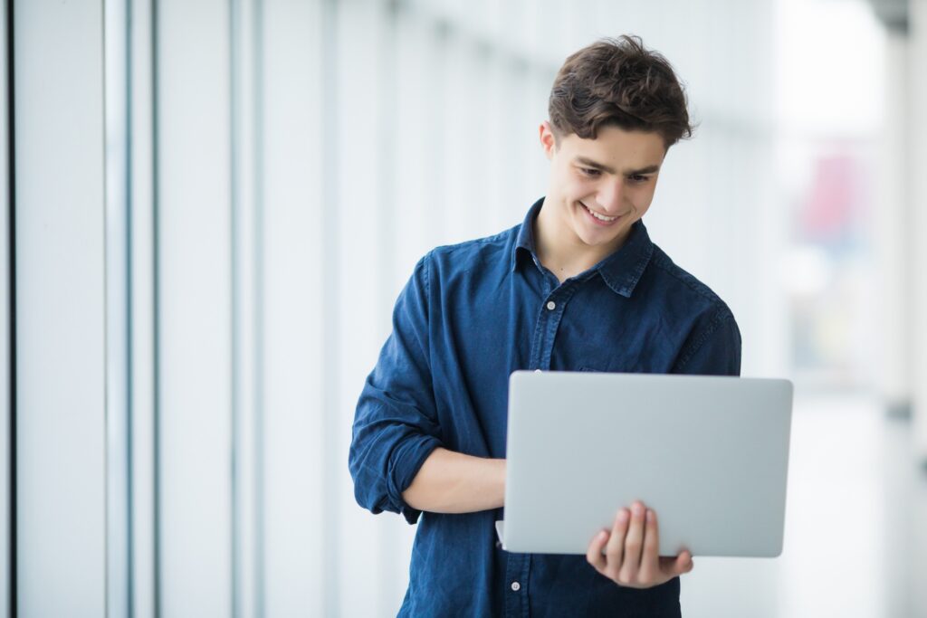 Boy holding a laptop and smiling next to windows.