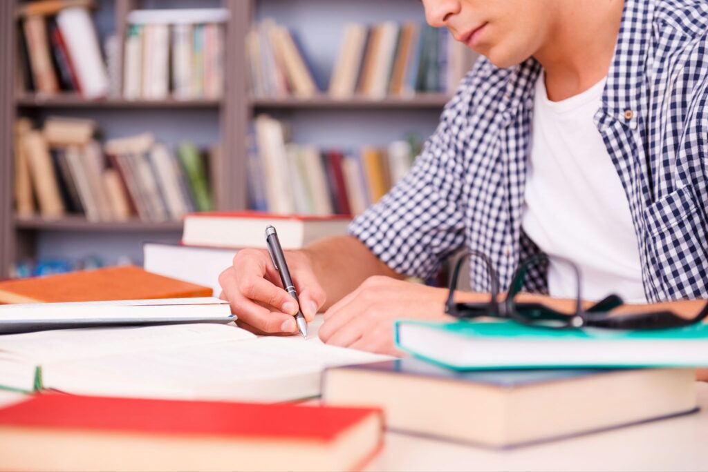 Male student using a pen next to textbooks, a pair of glasses, and a bookshelf in the background.