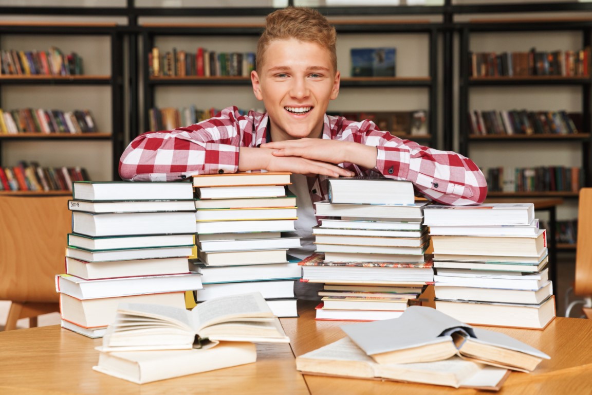 Smiling teenage boy sitting at the library table with big stacks of books.