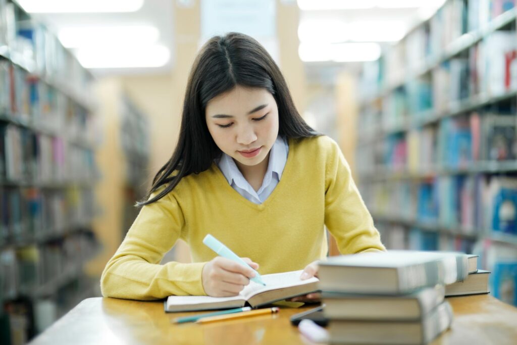 Girl studying in the library and holding a highlighter next to a pencil and a stack of books while sitting at a desk.